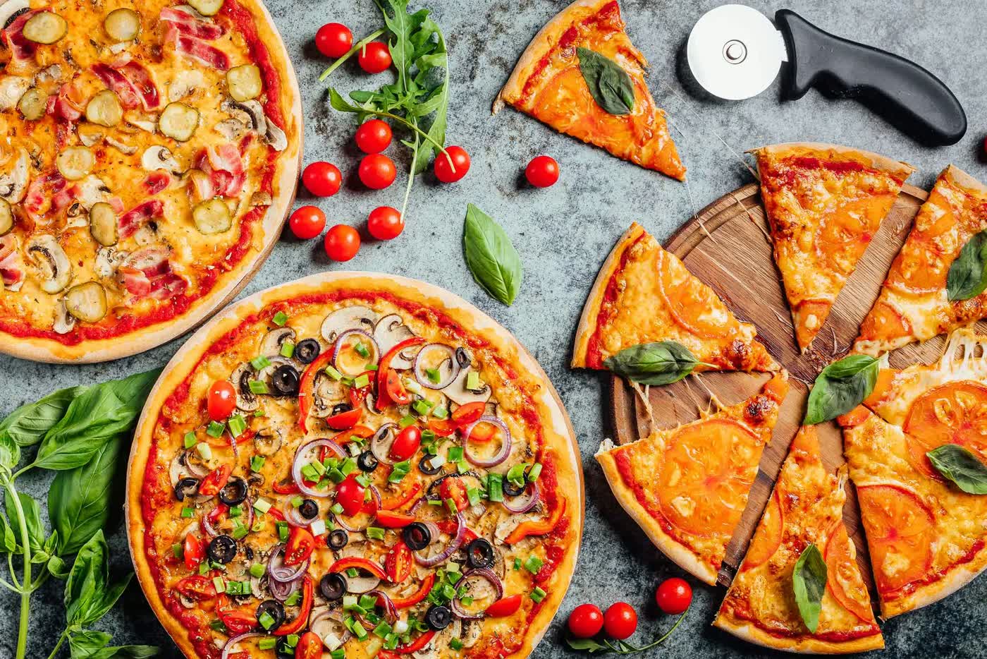 Drie pizza&rsquo;s van bovenaf op een grijze steen: een met spek, champignon en augurk, een met paprika, rode ui en olijf, en een in punten gesneden pizza met tomaat op een houten plank, met cherrytomaatjes, basilicum en een pizzasnijder eromheen.
