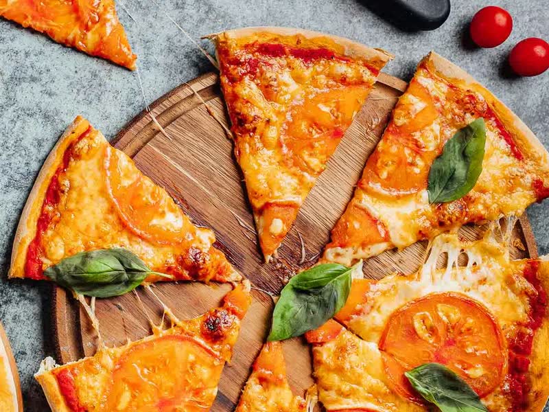 Een in punten gesneden pizza met tomaat en basilicum op een houten plank, met cherrytomaatjes en een pizzasnijder ernaast.